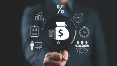 A person in a suit holding a magnifying glass highlighting a money bag icon surrounded by financial icons