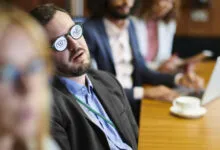 A tired businessman wearing novelty glasses with open eyes drawn on lenses during a meeting.