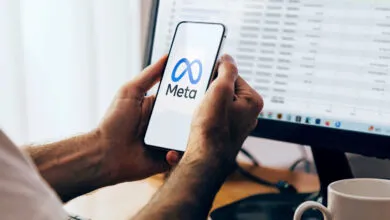 Man holding a smartphone displaying the blue Meta logo, with a computer monitor showing a dashboard behind