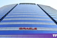 Low-angle view of the Oracle building facade featuring blue glass and red signage against the sky.