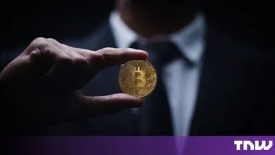 A person in a suit holding a shiny gold Bitcoin cryptocurrency token between fingers.