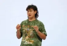 A man wearing a green deer graphic t-shirt speaks against a light blue background.