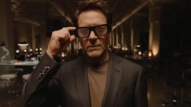 Robert Downey Jr. adjusts thick black smart glasses while wearing a dark blazer in a dimly lit room.