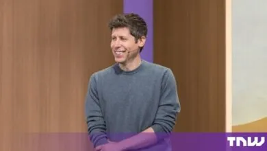 Sam Altman smiling on stage wearing a gray sweater against a warm wooden background
