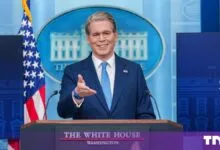 Scott Bessent smiling at the White House press briefing podium with an American flag behind him.
