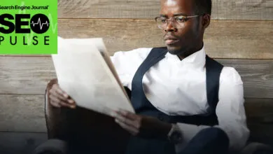 A man in glasses and vest reads a newspaper against a rustic wooden wall.