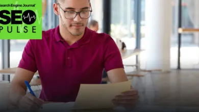 A young man wearing glasses and a maroon polo shirt reviews documents at a desk.