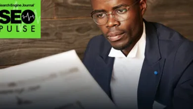 A Black man wearing glasses and a suit reads a newspaper in front of a wooden wall.