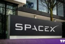 A black SpaceX logo sign standing outside a modern office building with glass windows.