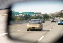 Golden Tesla Cybercab autonomous vehicle driving on a multi-lane highway, viewed through a rearview mirror.
