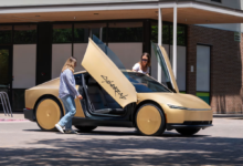 Two women stand beside a gold Tesla Cybercab with its futuristic butterfly doors open wide.