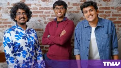 Three smiling young men standing in front of an exposed brick wall, dressed in casual clothing.