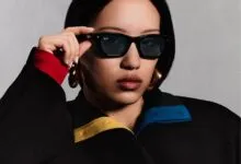Close-up of a stylish woman adjusting black Ray-Ban sunglasses wearing a black jacket with color accents.