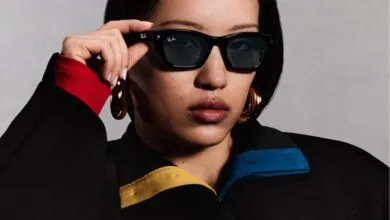 Close-up of a stylish woman adjusting black Ray-Ban sunglasses wearing a black jacket with color accents.