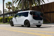 A white Waymo autonomous vehicle drives down a sunny street lined with palm trees.