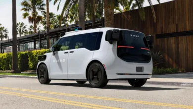A white Waymo autonomous vehicle drives down a sunny street lined with palm trees.